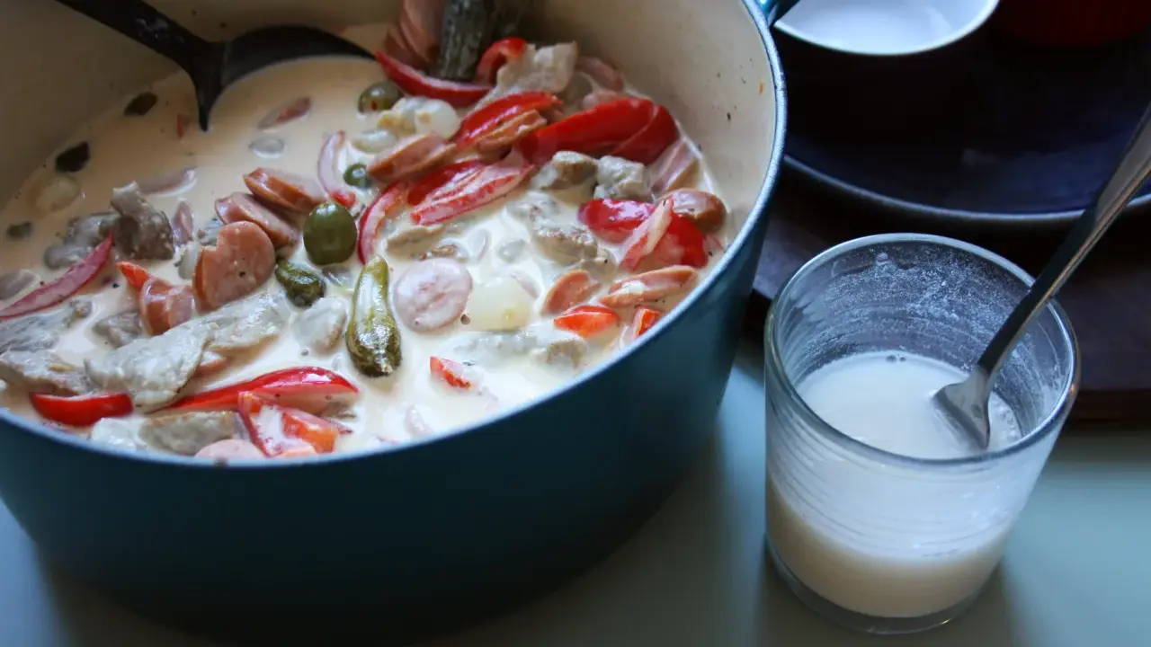 En krämig kreolsk gryta med korv, paprika, oliver och pickles i en blå gryta. En skål med mjölk står bredvid.