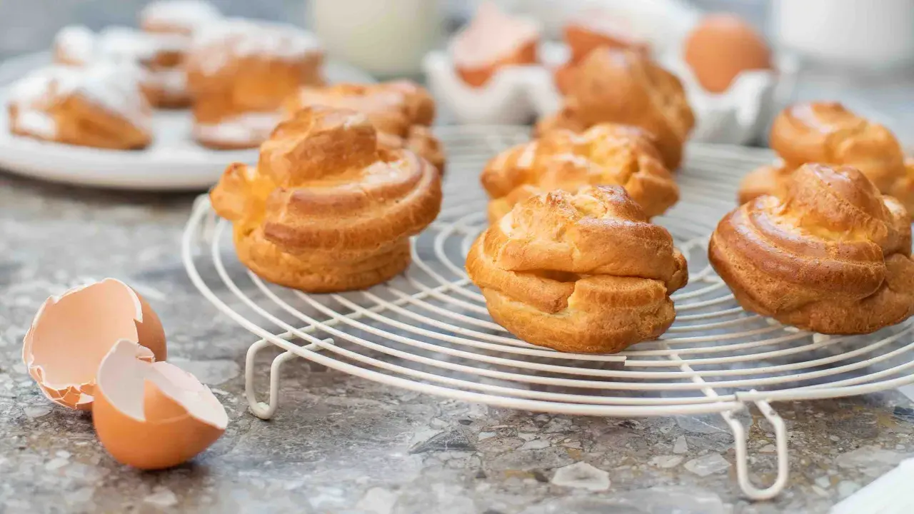 Nybakade petit choux på ett vitt galler, med äggskal och ägg i bakgrunden. Perfekt för fyllning!