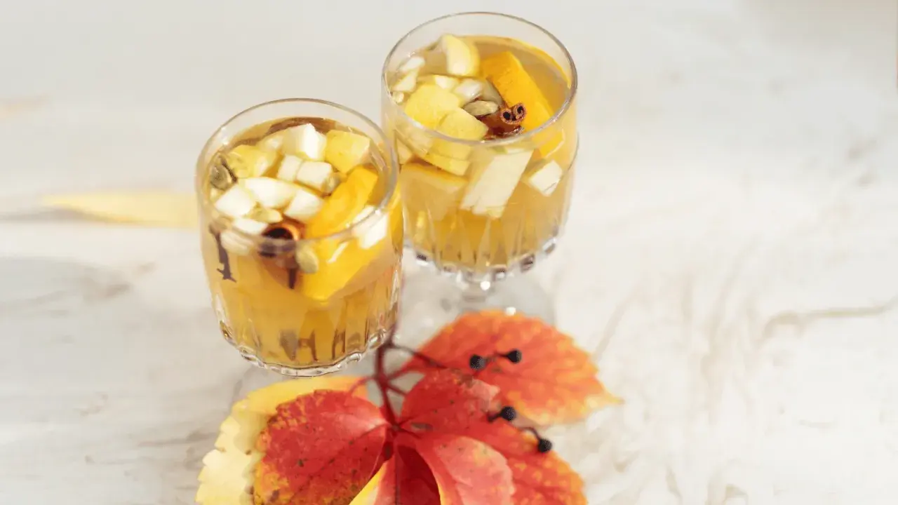 Två glas med gyllene vit sangria, fyllda med fruktbitar och kryddor, står på ett vitt bord. Höstlöv i rött och gult ligger framför.