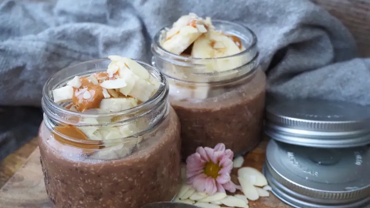 Två burkar med choklad overnight oats, toppade med banan, mandelflagor och en sked. Perfekt för en snabb frukost.