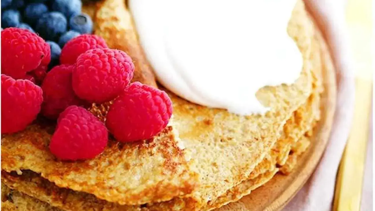 En stapel pannkakor med havregryn, toppade med grädde, hallon och blåbär. En gyllene gaffel ligger bredvid.