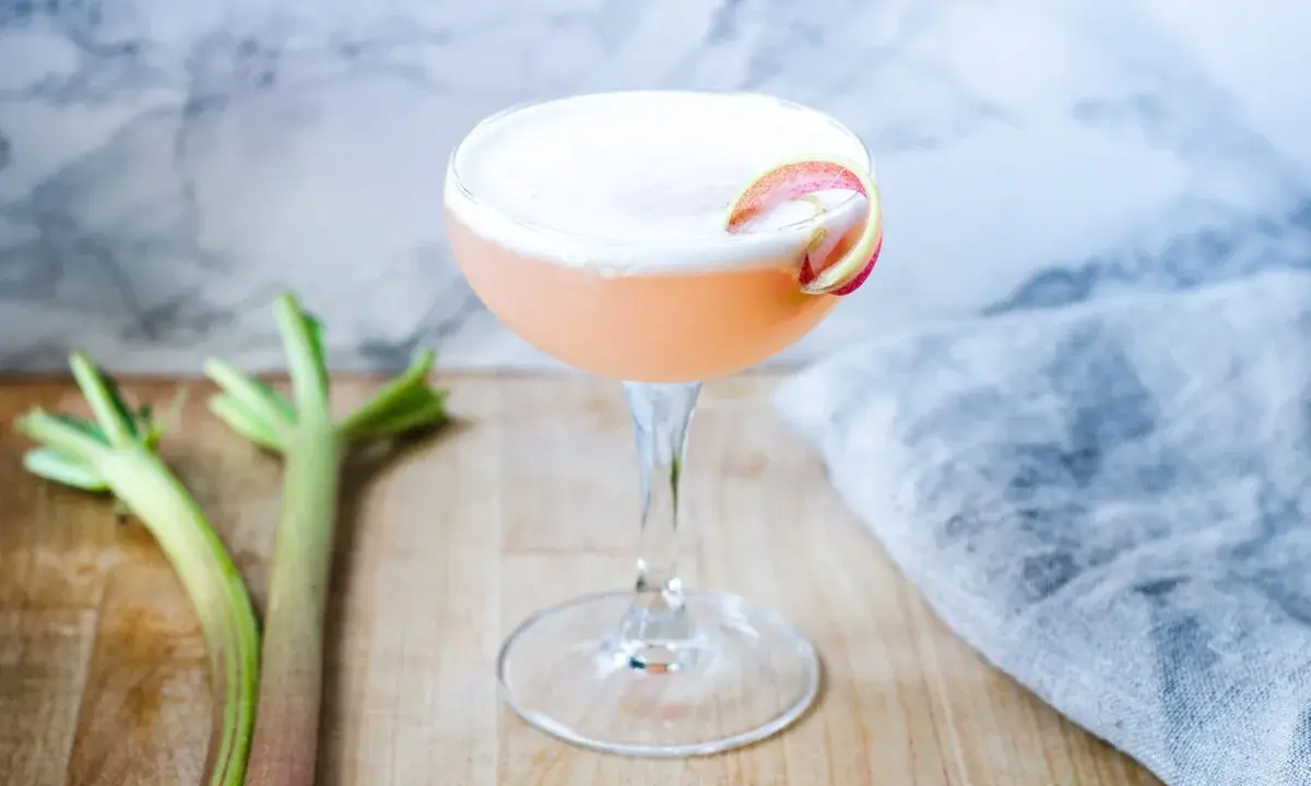 En rosa drink med rabarbersirap och gin, garnerad med en rabarberskiva. Bredvid syns färska rabarberstjälkar.
