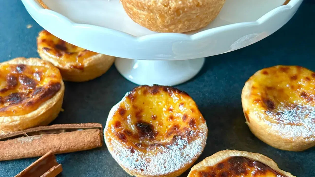 Flera gyllene pastel de nata, pudrade med florsocker, ligger utspridda på en mörk yta. Kanelstänger ligger bredvid.