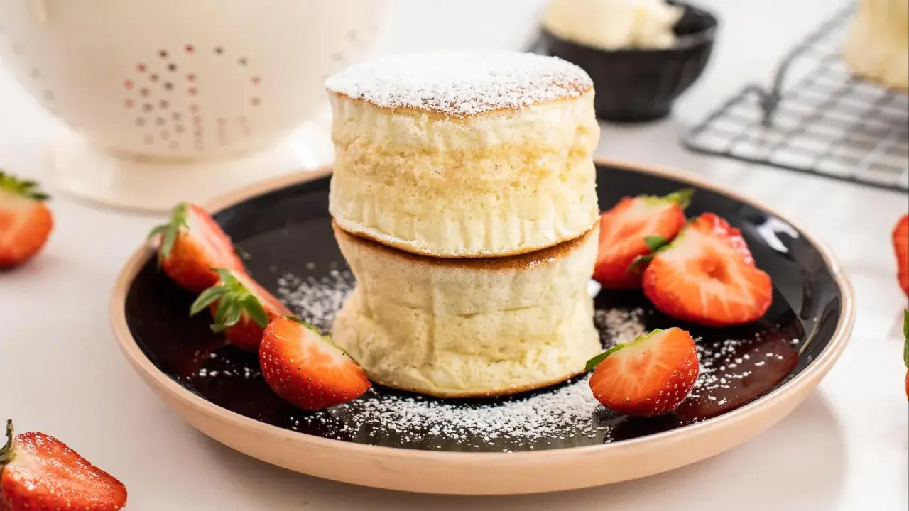 Två fluffiga japanska pannkakor staplade på en svart tallrik, garnerade med florsocker och färska jordgubbar.