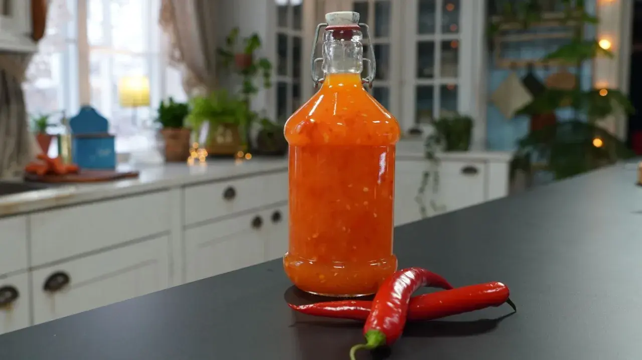 En flaska med hemgjord sweet chili sås står på en mörk bänk, med två röda chilifrukter bredvid.