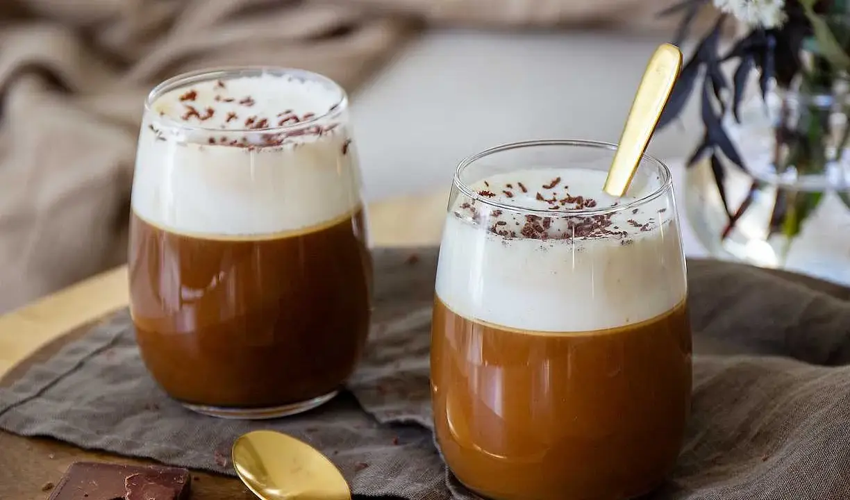 Två glas med krämig kaffe Karlsson, toppad med vispad grädde och chokladströssel. En gyllene sked vilar i det ena glaset.