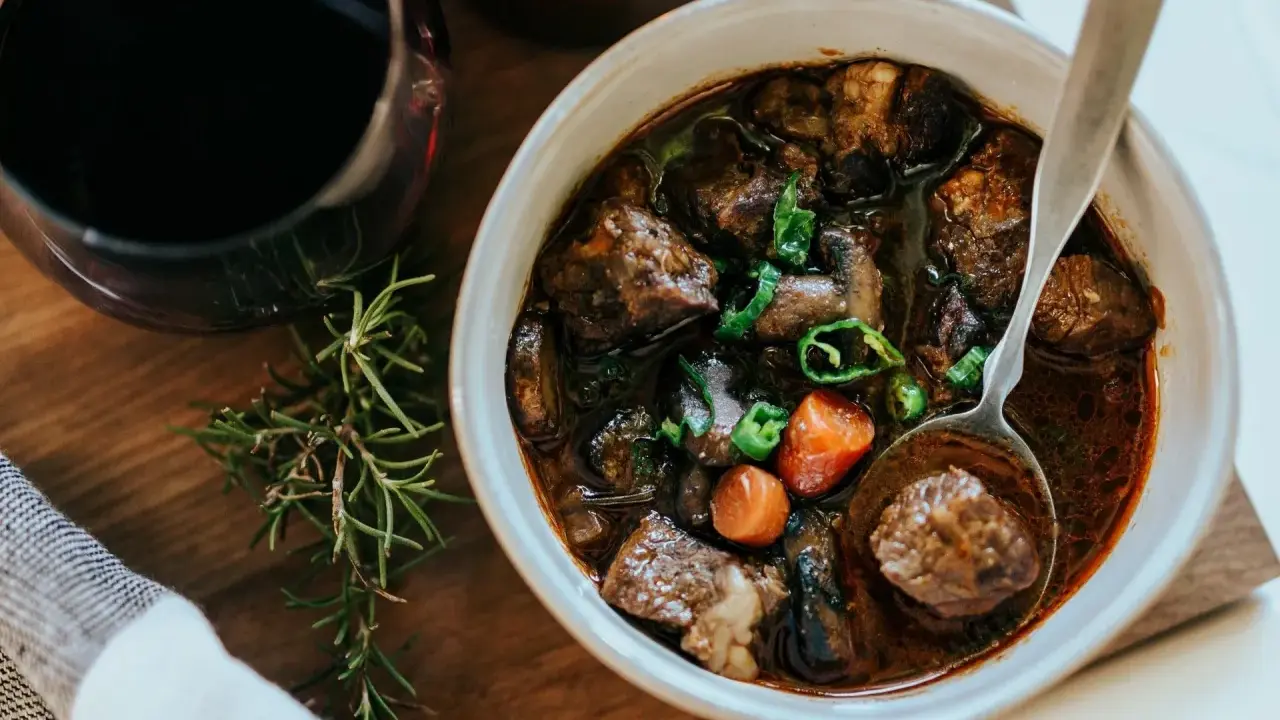 En skål med mustig boeuf bourguignon, perfekt för ett mysigt recept. Serveras med en sked och en kvist rosmarin.