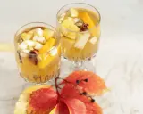 Två glas med gyllene vit sangria, fyllda med fruktbitar och kryddor, står på ett vitt bord. Höstlöv i rött och gult ligger framför.