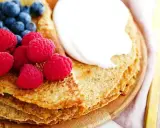 En stapel pannkakor med havregryn, toppade med grädde, hallon och blåbär. En gyllene gaffel ligger bredvid.