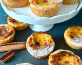 Flera gyllene pastel de nata, pudrade med florsocker, ligger utspridda på en mörk yta. Kanelstänger ligger bredvid.