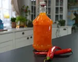En flaska med hemgjord sweet chili sås står på en mörk bänk, med två röda chilifrukter bredvid.