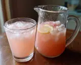 En glas och en karaff med rosa rabarbersirap drink fylld med is och citronskivor.