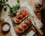 Två skivor rostat bröd med tomater och basilika, redo att avnjutas som ett enkelt bruschetta recept. En flaska olivolja och en skål flingsalt finns i närheten.