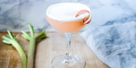 En rosa drink med rabarbersirap och gin, garnerad med en rabarberskiva. Bredvid syns färska rabarberstjälkar.