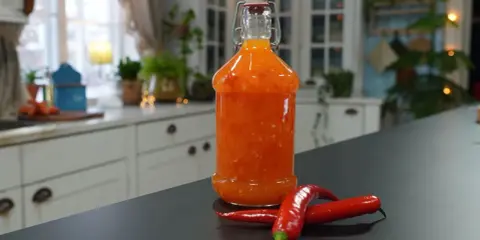 En flaska med hemgjord sweet chili sås står på en mörk bänk, med två röda chilifrukter bredvid.