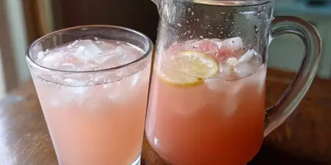 En glas och en karaff med rosa rabarbersirap drink fylld med is och citronskivor.