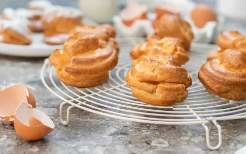 Nybakade petit choux på ett vitt galler, med äggskal och ägg i bakgrunden. Perfekt för fyllning!