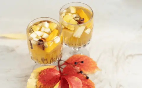 Två glas med gyllene vit sangria, fyllda med fruktbitar och kryddor, står på ett vitt bord. Höstlöv i rött och gult ligger framför.