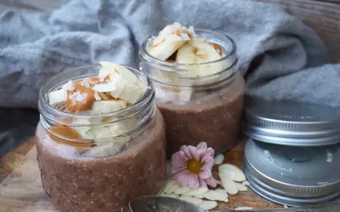 Två burkar med choklad overnight oats, toppade med banan, mandelflagor och en sked. Perfekt för en snabb frukost.