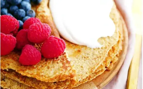 En stapel pannkakor med havregryn, toppade med grädde, hallon och blåbär. En gyllene gaffel ligger bredvid.