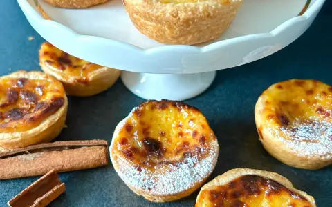 Flera gyllene pastel de nata, pudrade med florsocker, ligger utspridda på en mörk yta. Kanelstänger ligger bredvid.