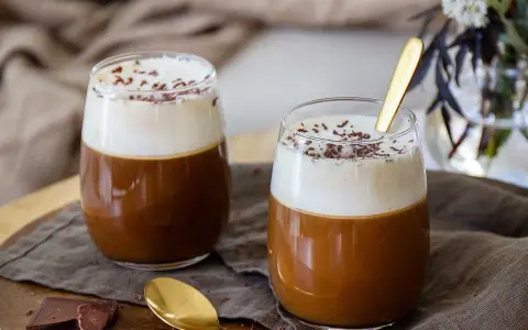 Två glas med krämig kaffe Karlsson, toppad med vispad grädde och chokladströssel. En gyllene sked vilar i det ena glaset.