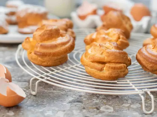 Nybakade petit choux på ett vitt galler, med äggskal och ägg i bakgrunden. Perfekt för fyllning!