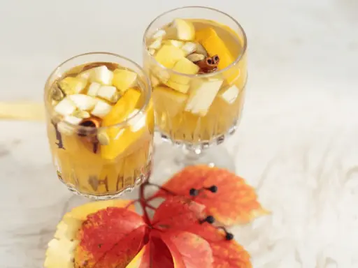 Två glas med gyllene vit sangria, fyllda med fruktbitar och kryddor, står på ett vitt bord. Höstlöv i rött och gult ligger framför.