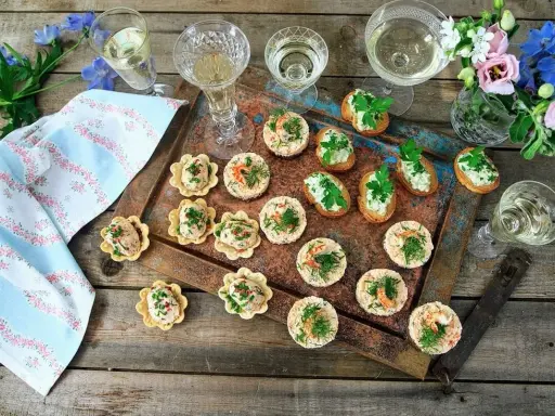 Små, läckra tilltugg till bubbel på ett rustikt fat, med glas fyllda med vitt vin och blommor i bakgrunden.