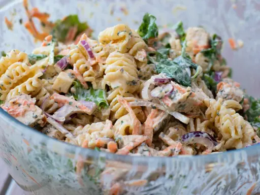 En stor skål med krämig pastasallad med kyckling, morötter, rödlök och gröna blad. Perfekt för en somrig picknick.