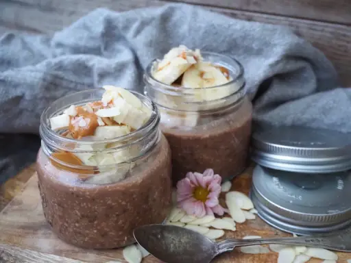 Två burkar med choklad overnight oats, toppade med banan, mandelflagor och en sked. Perfekt för en snabb frukost.
