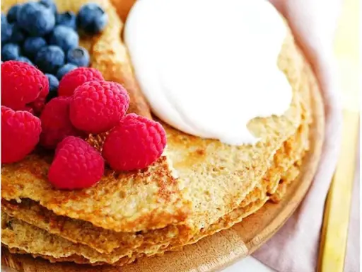 En stapel pannkakor med havregryn, toppade med grädde, hallon och blåbär. En gyllene gaffel ligger bredvid.