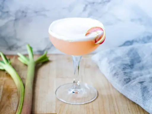 En rosa drink med rabarbersirap och gin, garnerad med en rabarberskiva. Bredvid syns färska rabarberstjälkar.