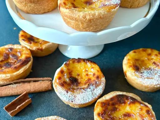 Flera gyllene pastel de nata, pudrade med florsocker, ligger utspridda på en mörk yta. Kanelstänger ligger bredvid.