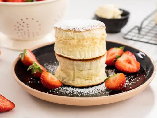 Två fluffiga japanska pannkakor staplade på en svart tallrik, garnerade med florsocker och färska jordgubbar.