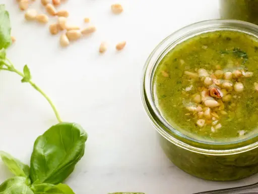 Färska basilikablad och pinjenötter bredvid en burk med hemlagad pesto genovese recept.