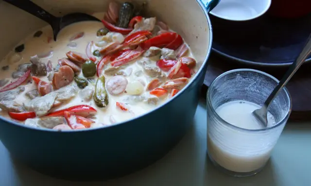 En krämig kreolsk gryta med korv, paprika, oliver och pickles i en blå gryta. En skål med mjölk står bredvid.