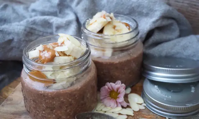 Två burkar med choklad overnight oats, toppade med banan, mandelflagor och en sked. Perfekt för en snabb frukost.