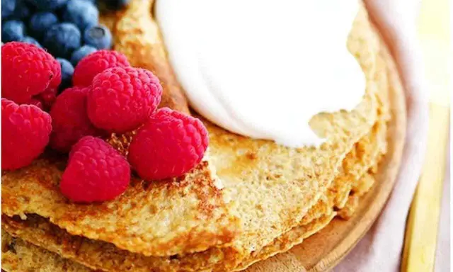 En stapel pannkakor med havregryn, toppade med grädde, hallon och blåbär. En gyllene gaffel ligger bredvid.