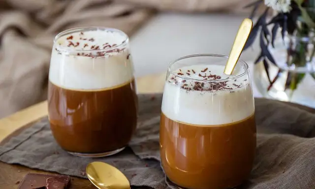 Två glas med krämig kaffe Karlsson, toppad med vispad grädde och chokladströssel. En gyllene sked vilar i det ena glaset.