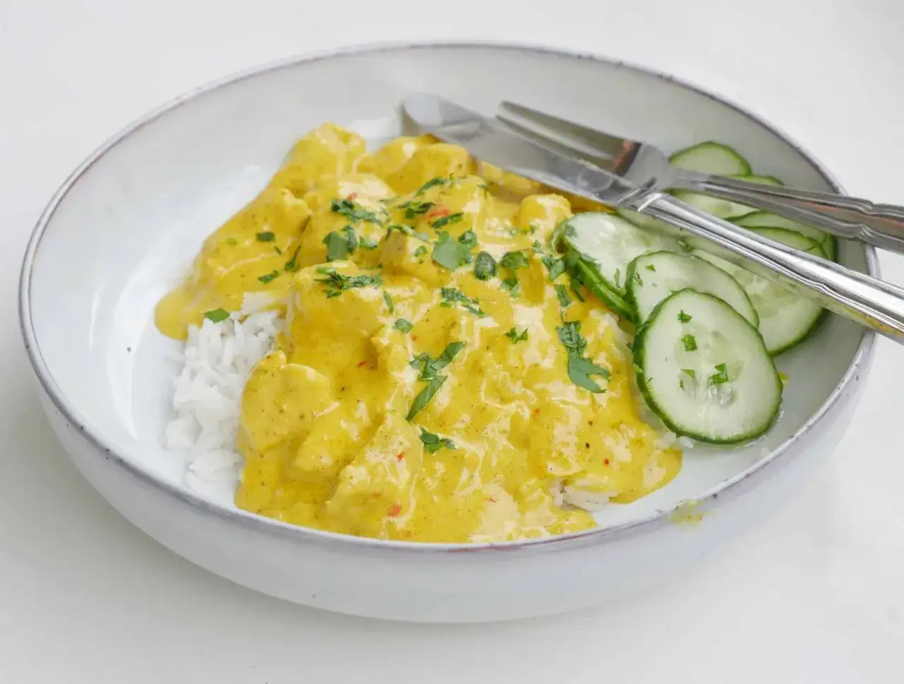 En skål med krämig kyckling curry över ris, garnerad med färsk persilja och serverad med gurkskivor. En härlig måltid!