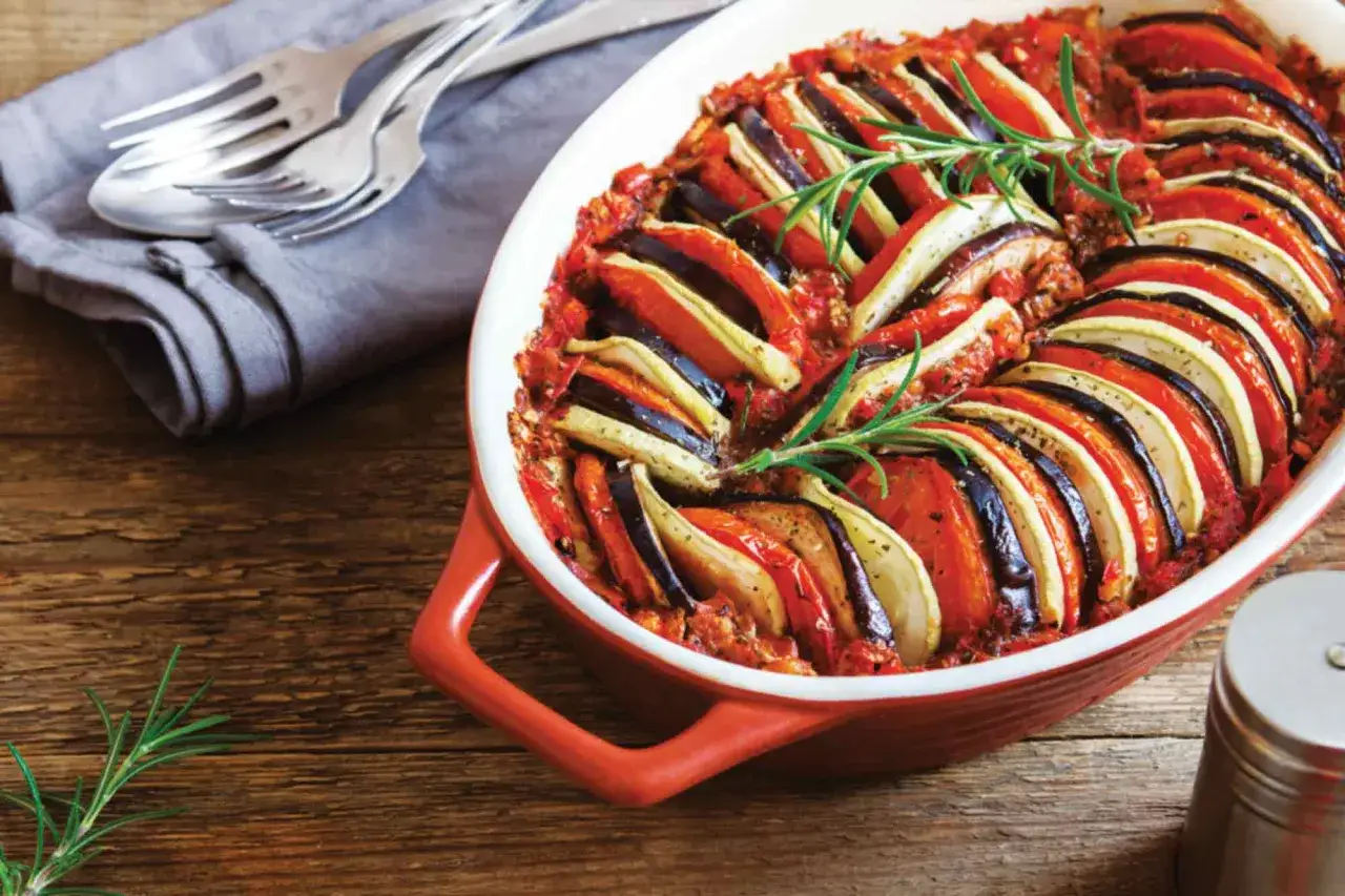 Enkel lyxig middag: vackert upplagd ratatouille i ugnsform, garnerad med rosmarin. Serveras med bestick på en grå duk.