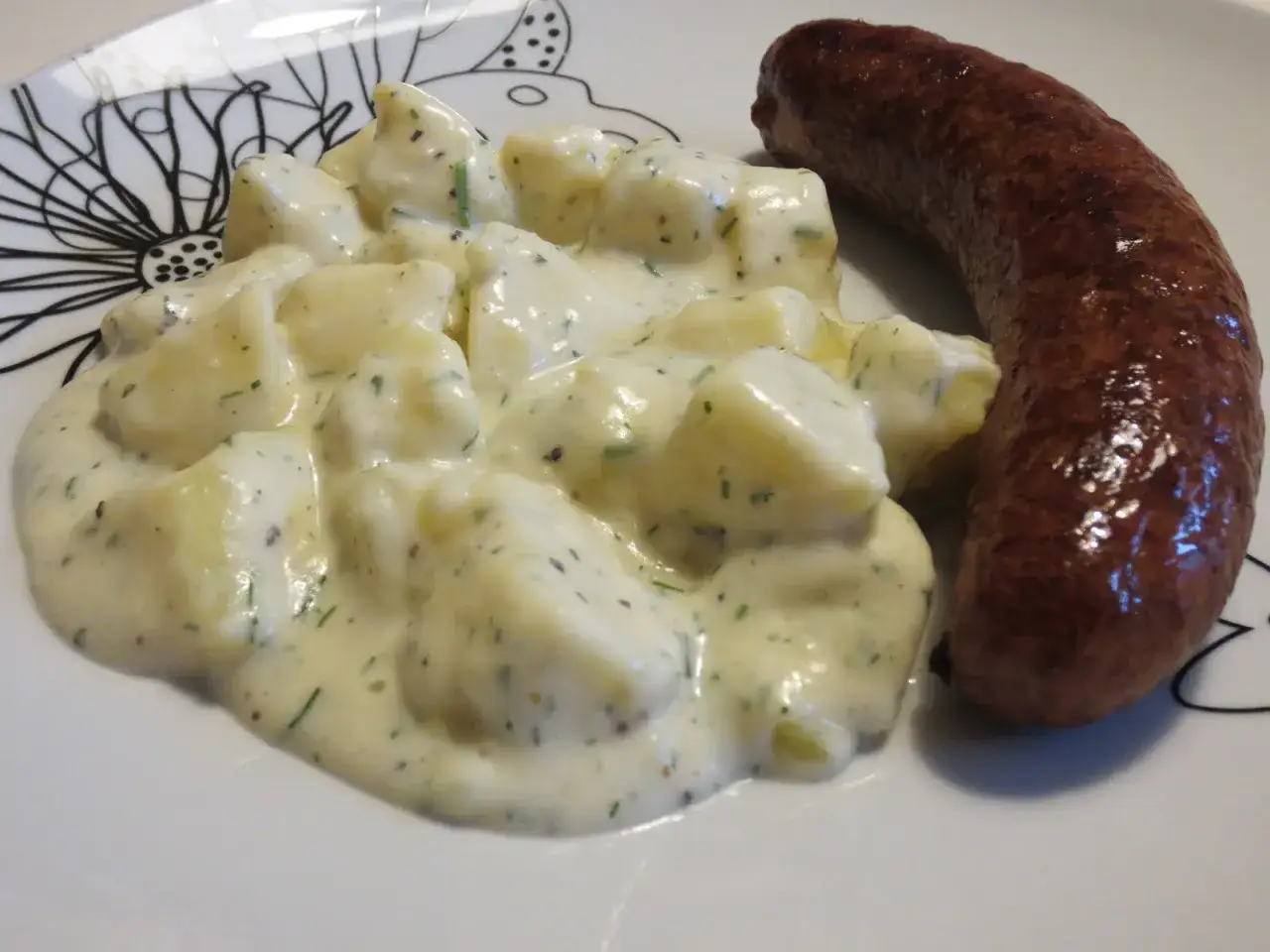 En tallrik med stuvad potatis och en korv. Potatisen är krämig och har gröna örter i sig.