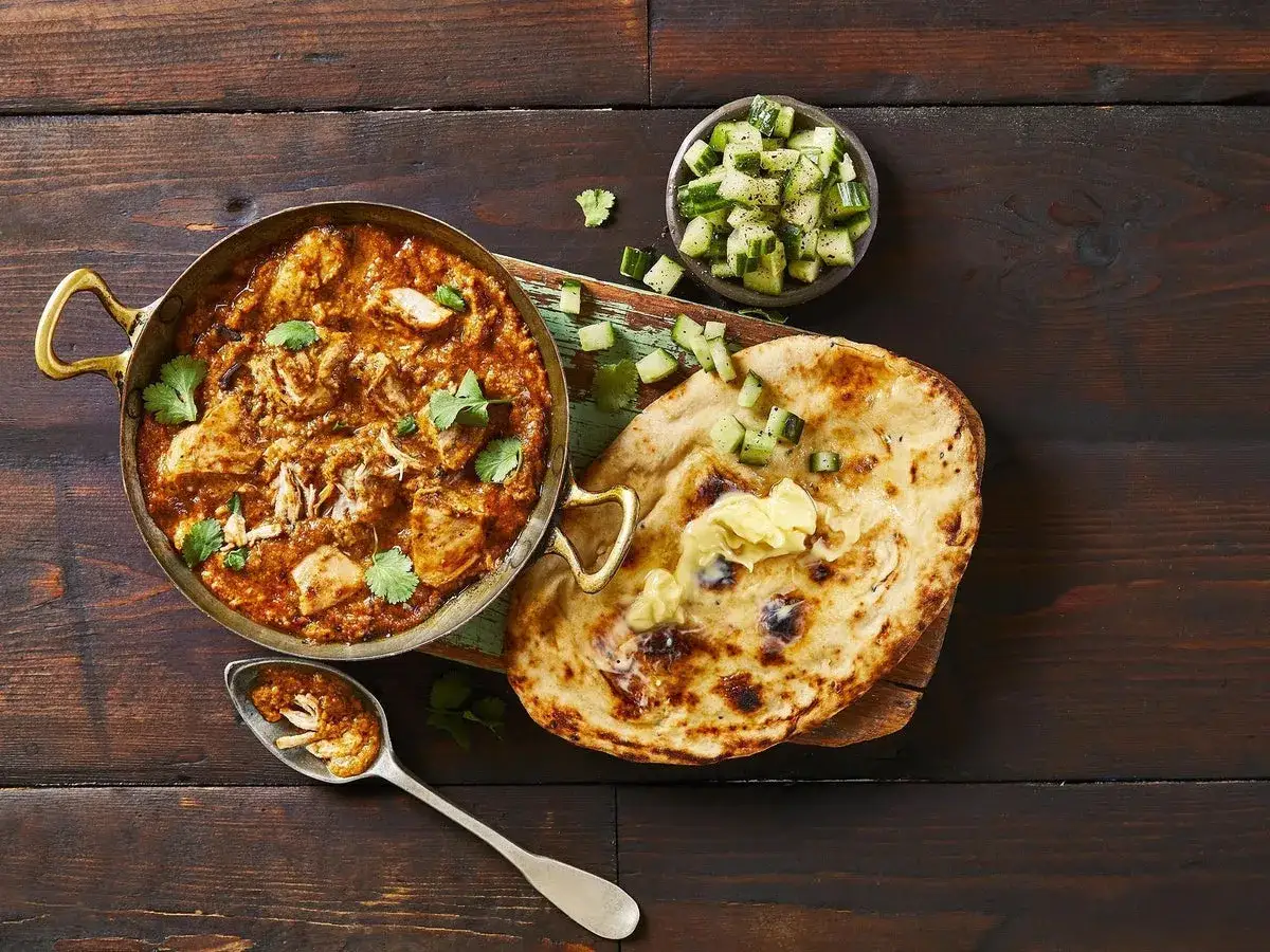 En skål med krämig butter chicken, serverad med naanbröd, gurksallad och en sked full av smak.