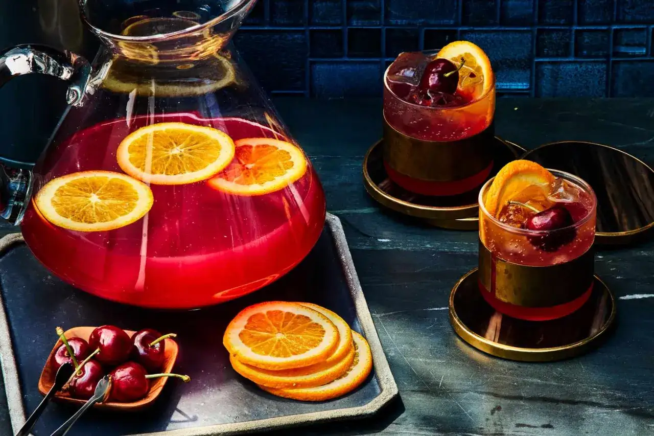 En karaff med rosa drink och apelsinskivor, bredvid två glas med enkla drinkar med ljus rom, körsbär och apelsin.
