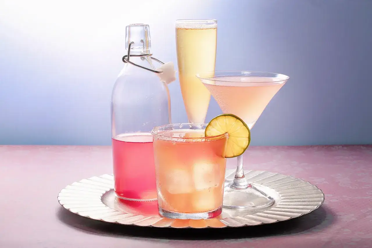 En flaska med rosa rabarbersirap och tre drinkar, en med lime, på ett silverfärgat fat.