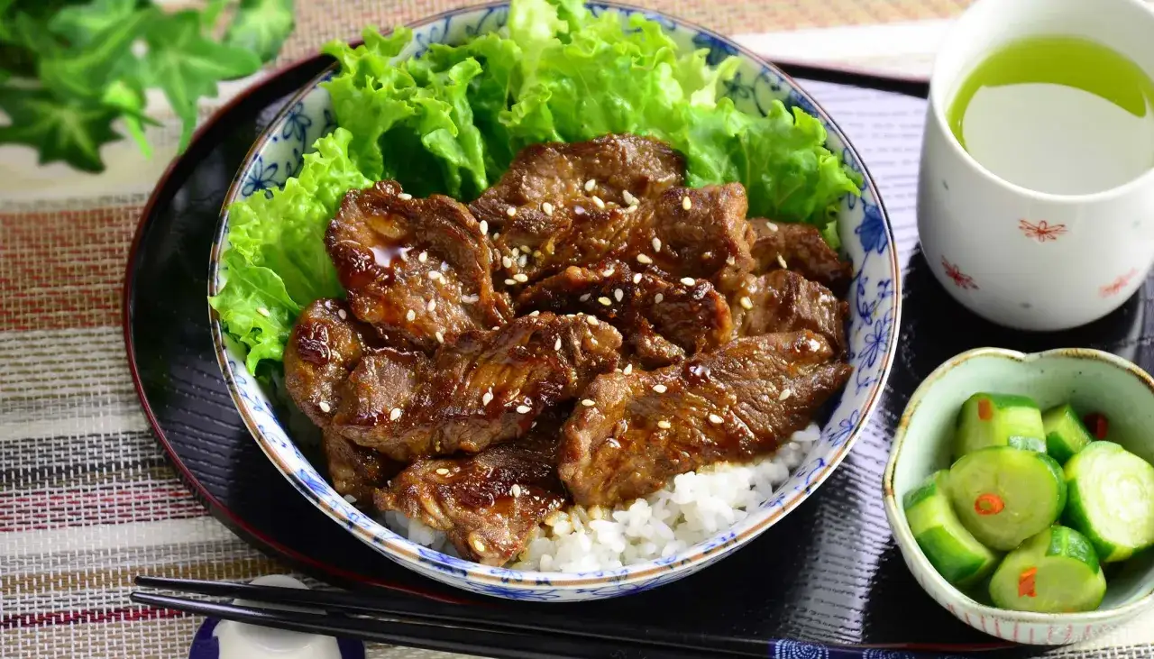 En skål med ris toppad med saftigt yakiniku-kött, garnerad med sesamfrön och sallad. Serveras med inlagd gurka och grönt te. Ett perfekt yakiniku recept!
