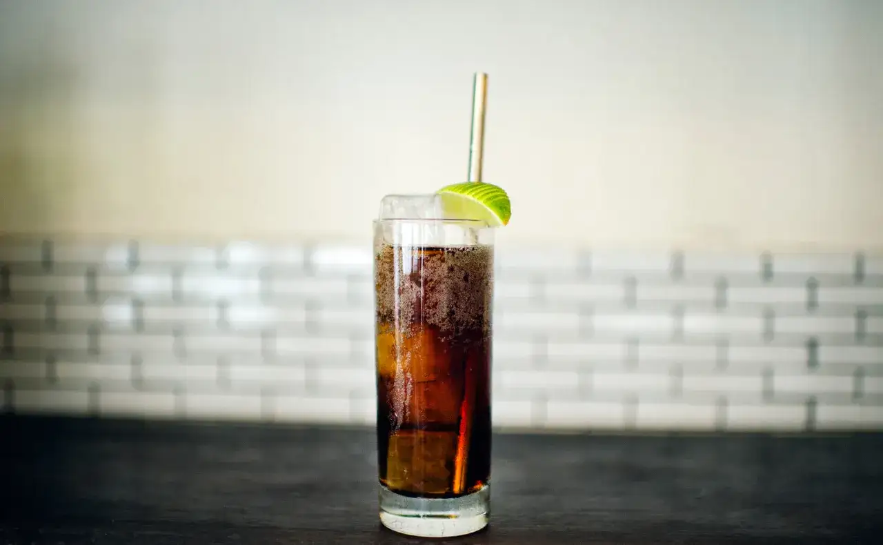Ett glas med en cuba libre drink, is, lime och ett sugrör.