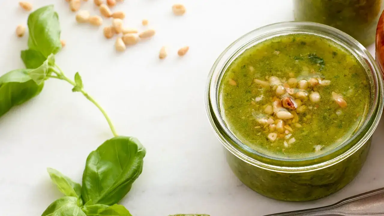 Färska basilikablad och pinjenötter bredvid en burk med hemlagad pesto genovese recept.