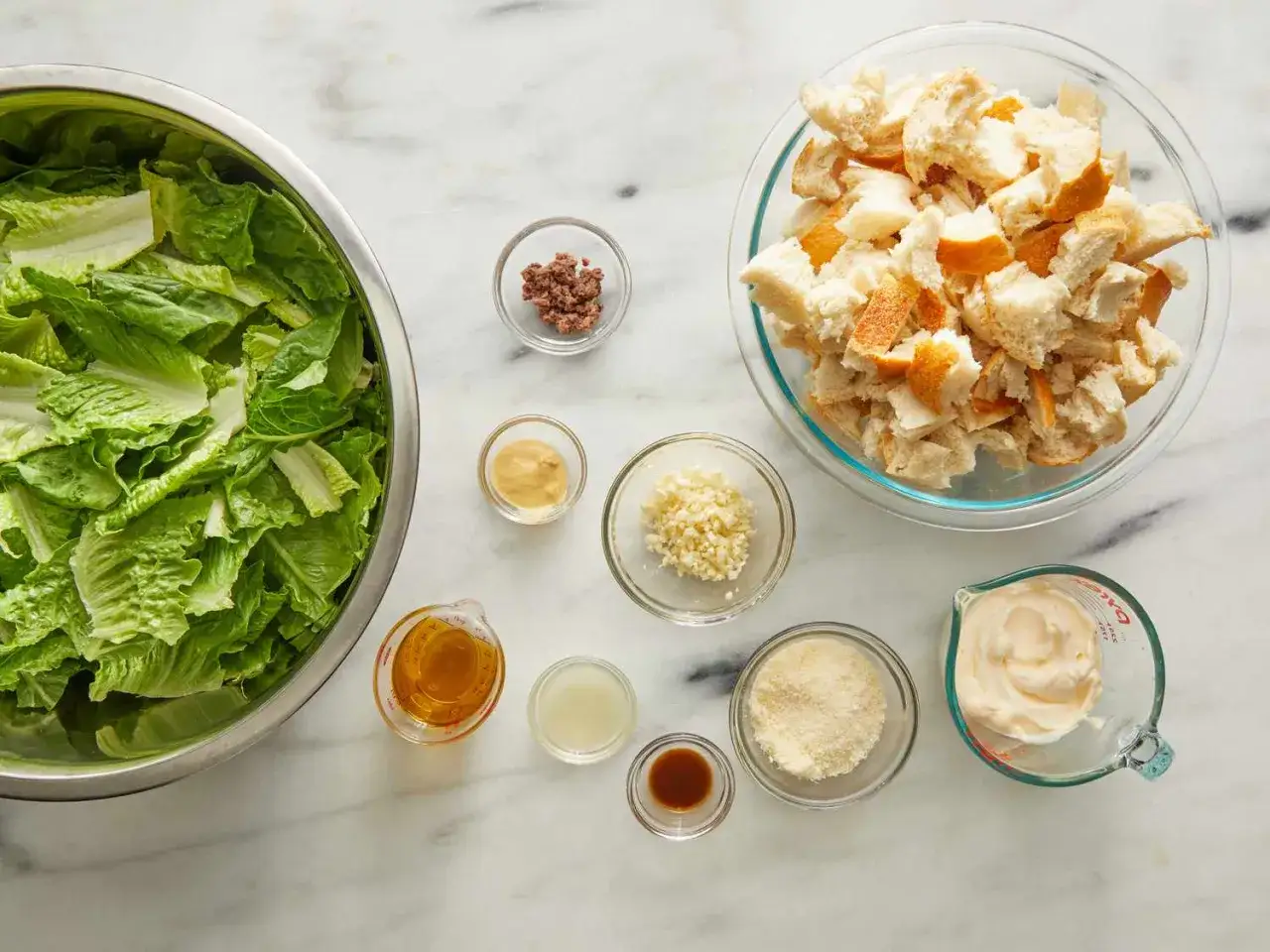 Alla ingredienser för en klassisk caesarsallad, inklusive romansallat, krutonger och skålar med vitlök, parmesan, ansjovis och en krämig caesarsallad dressing.